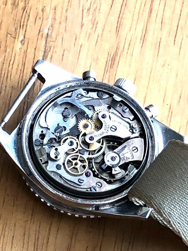 May include: Close-up of a vintage wristwatch's intricate internal mechanism. The watch features numerous small gears, jewels, and other components. The watch has a silver case and a green fabric strap. The watch is on a wooden surface.