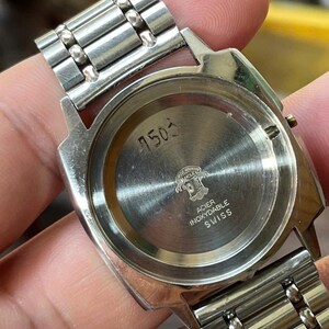以下が含まれることがあります： シルバーカラーの腕時計のクローズアップ。金属製のバンドと丸い文字盤が特徴です。時計の裏側には、数字の「7503」、ロゴ、そして「ACIER INOXYDABLE SWISS」の文字が表示されています。