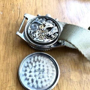 May include: A vintage silver-toned wristwatch with the back removed, revealing the intricate internal mechanisms. The watch has a light-colored canvas strap. The watch's back is also visible, with the text "ULYSSE NARDIN LOCLE SUISSE" and the number "432234".