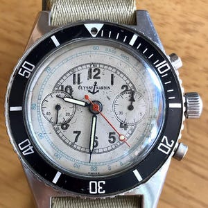 May include: Vintage Ulysse Nardin chronograph watch with a silver dial, black bezel, and beige canvas strap. The watch features multiple sub-dials, a red second hand, and luminous markers. The case is stainless steel.