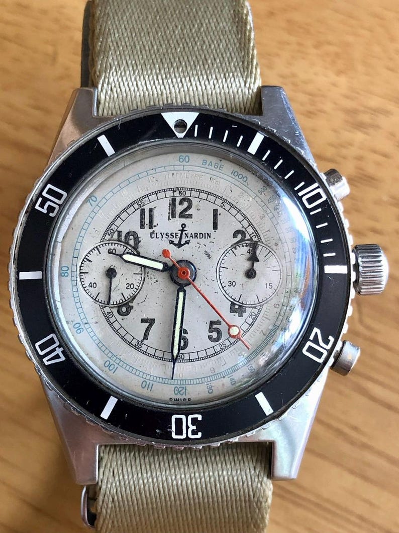 May include: Vintage Ulysse Nardin chronograph watch with a silver dial, black bezel, and beige canvas strap. The watch features multiple sub-dials, a red second hand, and luminous markers. The case is stainless steel.