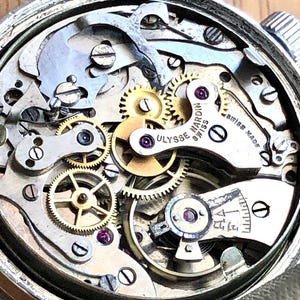 May include: Close-up of a vintage Ulysse Nardin Swiss Made watch movement. The intricate mechanism features numerous gears, screws, and jewels in a silver-toned case. The watch's internal components are visible, showcasing the precision of the craftsmanship.
