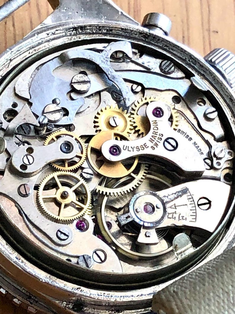May include: Close-up of a vintage Ulysse Nardin Swiss Made watch movement. The intricate mechanism features numerous gears, screws, and jewels in a silver-toned case. The watch's internal components are visible, showcasing the precision of the craftsmanship.