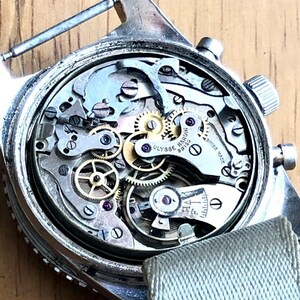 May include: Close-up of a vintage silver wristwatch with the back removed, revealing intricate mechanical components. The watch mechanism includes numerous gears, springs, and jewels. A light green fabric strap is attached. The watch face is marked with the brand name "Ulysse Nardin".