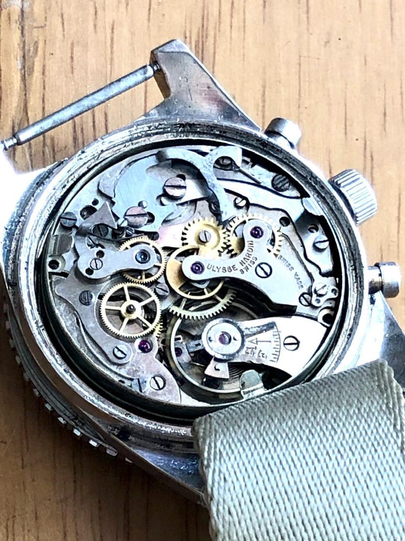 May include: Close-up of a vintage silver wristwatch with the back removed, revealing intricate mechanical components. The watch mechanism includes numerous gears, springs, and jewels. A light green fabric strap is attached. The watch face is marked with the brand name "Ulysse Nardin".