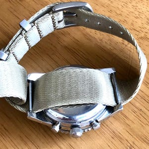 May include: A silver-toned wristwatch with a light green canvas band. The watch face is not visible, but the side view shows the crown and buttons. The band is secured with a silver buckle. The watch is on a wooden surface.
