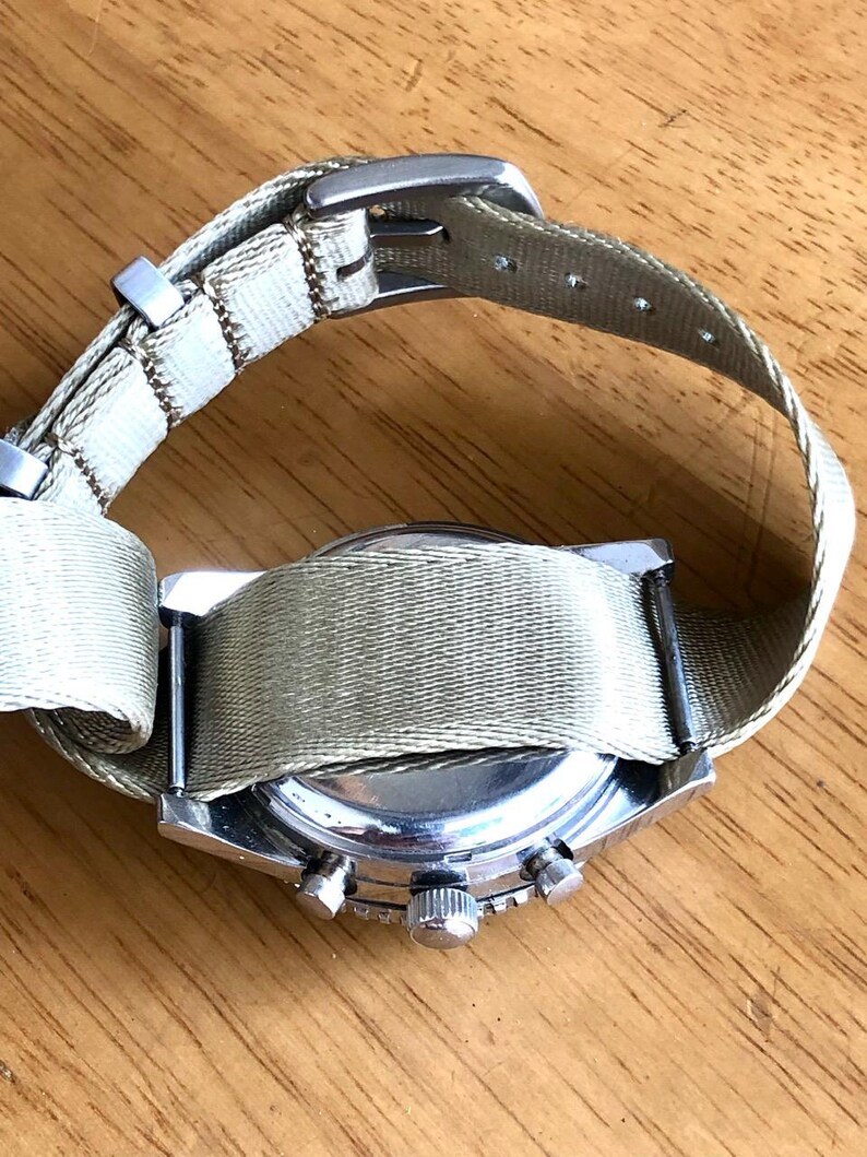 May include: A silver-toned wristwatch with a light green canvas band. The watch face is not visible, but the side view shows the crown and buttons. The band is secured with a silver buckle. The watch is on a wooden surface.