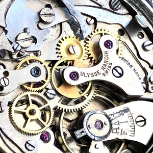May include: Close-up of a watch mechanism with intricate gears, screws, and a balance wheel. The watch movement features the text "ULYSSE NARDIN SWISS" and "SWISS MADE". The components are silver and gold in color, with red jewels.
