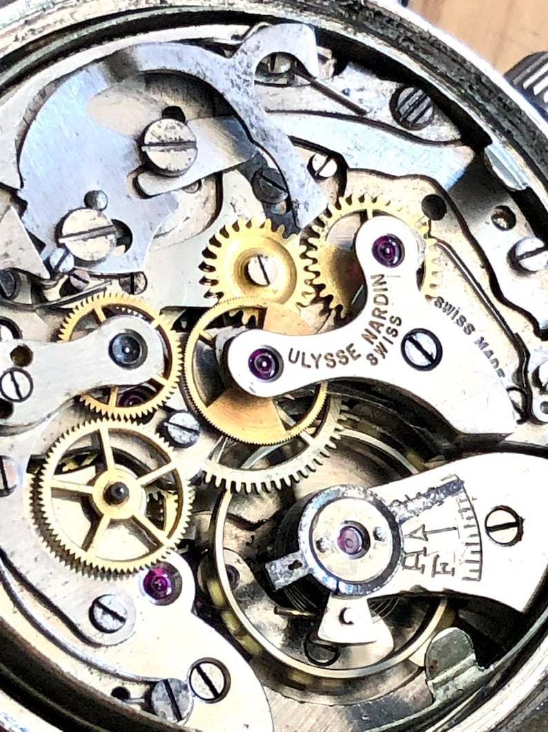 May include: Close-up of a watch mechanism with intricate gears, screws, and a balance wheel. The watch movement features the text "ULYSSE NARDIN SWISS" and "SWISS MADE". The components are silver and gold in color, with red jewels.