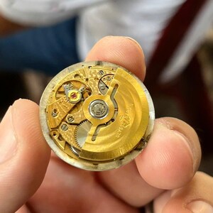 以下が含まれることがあります： 複雑な歯車と機構を備えた、金色の時計ムーブメントのクローズアップ。 時計のムーブメントは人の手で持たれています。 「ULYSSE NARDIN」の文字が見えます。