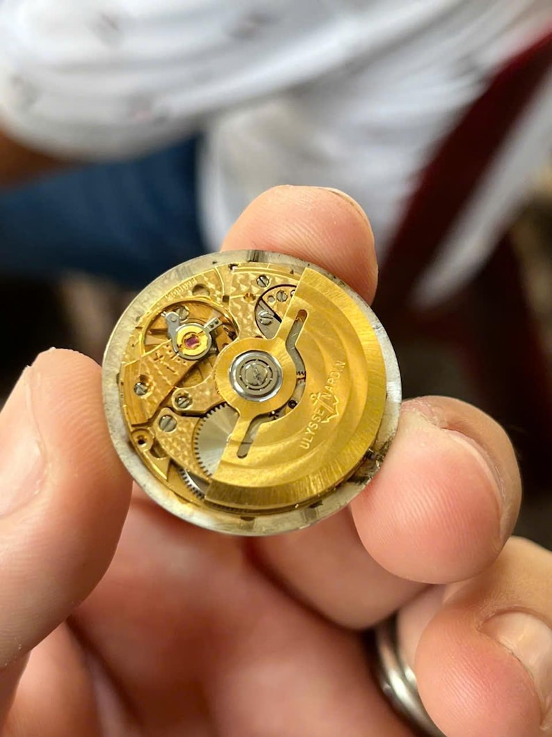 以下が含まれることがあります： 複雑な歯車と機構を備えた、金色の時計ムーブメントのクローズアップ。 時計のムーブメントは人の手で持たれています。 「ULYSSE NARDIN」の文字が見えます。