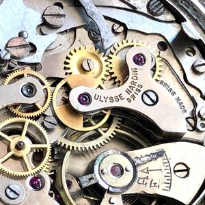 May include: Close-up of a watch mechanism with intricate gears and components. The image highlights the precision of the watchmaking, with visible text reading "Ulysse Nardin Swiss Made". The gears are gold and silver in color, with small ruby jewels.