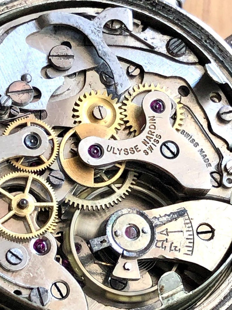 May include: Close-up of a watch mechanism with intricate gears and components. The image highlights the precision of the watchmaking, with visible text reading "Ulysse Nardin Swiss Made". The gears are gold and silver in color, with small ruby jewels.