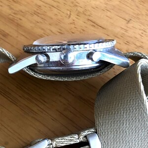 May include: A silver-toned wristwatch with a clear, domed crystal and a rotating bezel. The watch features two side buttons and a light green fabric band with a silver buckle. The watch is shown from a side profile.