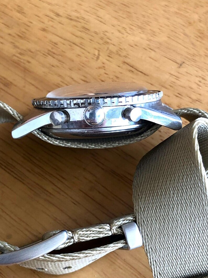 May include: A silver-toned wristwatch with a clear, domed crystal and a rotating bezel. The watch features two side buttons and a light green fabric band with a silver buckle. The watch is shown from a side profile.