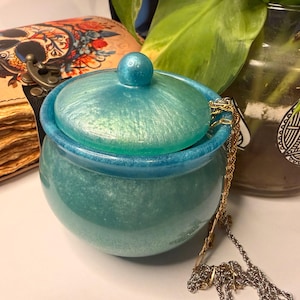 Può includere: Un barattolo rotondo color turchese con coperchio abbinato e un piccolo pomello. Il barattolo ha una finitura scintillante e iridescente. Una catena color oro pende dal barattolo. Un diario decorativo con un disegno a teschio è sullo sfondo.