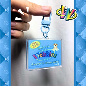 May include: A blue Webkinz keychain with a rectangular tag featuring a puppy illustration and the text "Webkinz." The keychain includes a blue clasp and a ring. The tag also has the text "Adopt a pet..." and "discover a virtual world!"