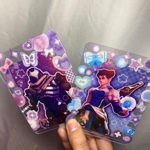 May include: Two rectangular clear plastic card holders decorated with purple and blue glittery stickers, including hearts, stars, and butterflies. Each holder contains a character card featuring a video game figure and the Overwatch logo.