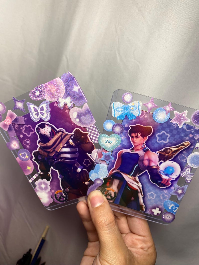 May include: Two rectangular clear plastic card holders decorated with purple and blue glittery stickers, including hearts, stars, and butterflies. Each holder contains a character card featuring a video game figure and the Overwatch logo.