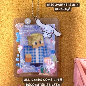 May include: A clear plastic toploader holding a card with a blocky, pixelated character wearing a blue plaid shirt. The toploader is decorated with various small stickers including a white cartoon dog, shells, and fish. Text reads: ALSO AVAILABLE AS A KEYCHAIN! and ALL CARDS COME WITH DECORATED STICKER TOPLOADERS.