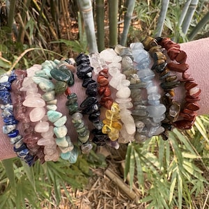 Può includere: Una collezione di braccialetti con schegge di pietre preziose in vari colori, tra cui blu, rosa, verde, nero, bianco, giallo e marrone. I braccialetti sono esposti su una superficie rosa, con uno sfondo naturale di bambù e vegetazione.