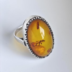 Op de afbeelding: Een zilveren ring met een grote, ovale, doorschijnende honingkleurige barnsteen. De barnsteen bevat zichtbare natuurlijke insluitingen en organisch materiaal. De steen is gezet in een decoratieve, gekartelde zilveren zetting met een gladde ringband.
