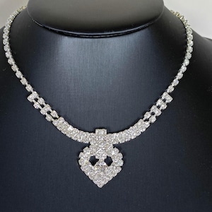 Op de afbeelding: Een zilverkleurige ketting met een doorlopende rij kleine, heldere, gefacetteerde strass-steentjes. In het midden van de ketting bevindt zich een hartvormige hanger bestaande uit meerdere heldere strass-steentjes, wat zorgt voor een heldere, reflecterende en sprankelende uitstraling.