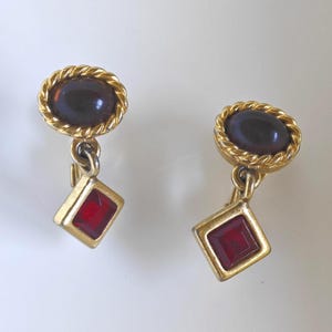 Può includere: Un paio di orecchini a clip color oro con una parte superiore rotonda caratterizzata da un cabochon ovale marrone scuro circondato da un bordo a corda intrecciata, collegato a un ciondolo a forma di rombo con una pietra quadrata rossa sfaccettata.