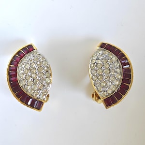 May include: A pair of gold-tone clip-on earrings featuring a semi-circular design. Each earring is adorned with a curved row of deep red baguette-cut stones along the outer edge and a cluster of clear round pavé-set crystals in the center.