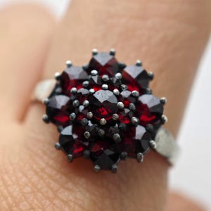Puede incluir: Anillo de plata con un grupo de piedras preciosas de color rojo intenso dispuestas en un patrón floral. Las piedras están facetadas, captando la luz y creando un efecto brillante. La banda del anillo es de plata pulida.
