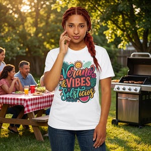 May include: White t-shirt with the text "Verano & Vibes Solsticios" in a colourful design featuring a sun, flowers, and a watermelon. The shirt is worn outdoors with a picnic table and grill in the background.