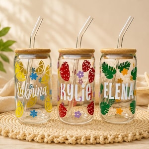 May include: Three clear glass can-shaped drinking glasses with bamboo lids and glass straws. Each glass is decorated with a different design and name: lemons and "Sabrina", strawberries and "Kylie", and tropical leaves and "Elena".