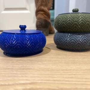 Puede incluir: Tres recipientes decorativos con tapa. Uno es azul vibrante, los otros están apilados, con una parte superior verde y una base gris. Cada uno tiene un patrón geométrico. Los recipientes están sobre una superficie de madera clara.