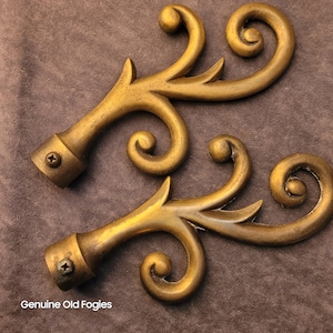 May include: Two antique gold-coloured curtain rod finials with a scrolled design. Each finial has a decorative, swirling pattern and a mounting base with a screw. The text "Genuine Old Fogies" is at the bottom.