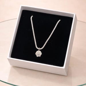 Peut inclure: Un collier en argent avec un grand pendentif rond en pierre précieuse transparente est présenté dans une boîte blanche à intérieur noir. La chaîne du collier est délicate et scintillante. La boîte repose sur une table en verre.