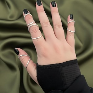 Könnte beinhalten: Ein Set aus sieben dünnen, glatten, silberfarbenen Metallringen, die an den Fingern getragen werden. Die Ringe sind schlichte, kreisförmige Bänder mit polierter Oberfläche, die an Daumen, Zeige-, Mittel- und Ringfinger sitzen und geschichtet wirken.