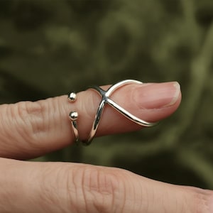 Puede incluir: Un anillo plateado con un diseño en forma de X y dos pequeños detalles redondos. El anillo se lleva en un dedo, mostrando su estilo moderno y minimalista. El diseño es simple pero llamativo, adecuado para el uso diario.