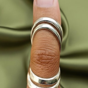 May include: Silver rings stacked on a finger. The rings are smooth and shiny, with a simple, minimalist design. The rings are worn on a finger, with the skin visible between the rings. The background is a solid olive green color.