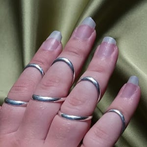 May include: A collection of six smooth, polished silver-toned metal rings worn on the fingers. Each ring features a simple, rounded band design with a reflective surface. The rings are positioned across multiple fingers, showcasing a uniform metallic finish.