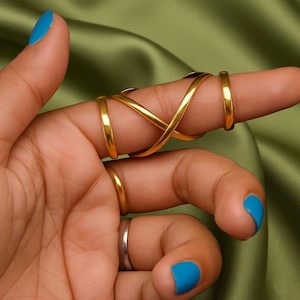 Può includere: Un set di anelli dorati. Il set include un anello con un design a simbolo dell'infinito e due fasce semplici. Gli anelli sono indossati su un dito con smalto blu. Lo sfondo è un tessuto verde e setoso.