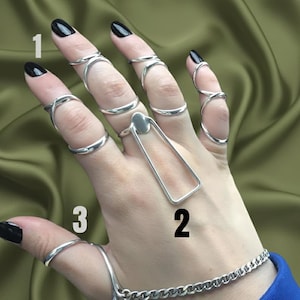 Op de afbeelding: Een hand met verschillende zilveren ringen. De ringen omvatten een vingerring, een lange rechthoekige ring en verschillende ringen die om de vingers wikkelen. Een zilveren kettingarmband is ook zichtbaar.