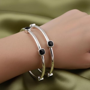 Op de afbeelding: Twee zilveren armbanden met vierkante zwarte edelstenen. De armbanden zijn gebogen en hebben een gepolijste afwerking. De edelstenen zijn gezet in een bezel setting. De achtergrond is een satijnen stof.