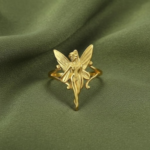 May include: A gold-colored ring featuring a fairy design. The fairy has outstretched wings and a slender body. The ring is set against a green background, highlighting the intricate details of the jewelry.