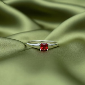 May include: A silver ring with a square, bright red gemstone. The ring is set against a backdrop of olive green fabric, creating a striking contrast. The gemstone is the focal point, catching the light and drawing the eye.