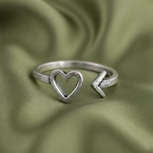 Puede incluir: Anillo plateado con un diseño en forma de corazón y una flecha que apunta hacia afuera. El anillo se encuentra sobre un fondo verde.