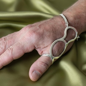 May include: A silver-toned bracelet with a unique design. The bracelet features a chain that connects to a large, openwork shape that wraps around the thumb and wrist. The jewelry is displayed against a green background.