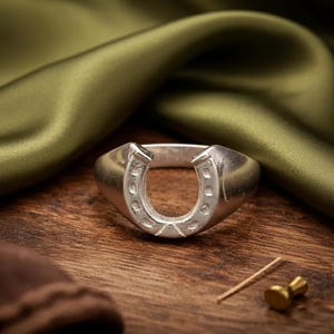 May include: A silver-colored ring with a horseshoe design. The ring is set against a wooden surface with a green fabric backdrop. The horseshoe is a classic symbol of luck and good fortune.