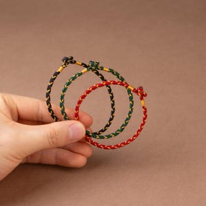 May include: Three braided bracelets in red, green, and navy blue with gold accents. The bracelets are held in a hand against a brown background. Each bracelet has a knotted closure.