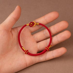 Puede incluir: Pulsera roja y dorada con diseño anudado. La pulsera presenta un cordón rojo vibrante con detalles dorados en los nudos y cierres. La pulsera se muestra sobre un fondo neutro.