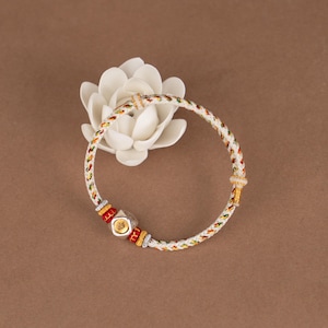 May include: A white and multicolored woven bracelet with gold accents and a silver charm. The bracelet features red, yellow, and green threads woven into the white cord. The charm has a gold border and a silver center. The bracelet is displayed on a brown surface.